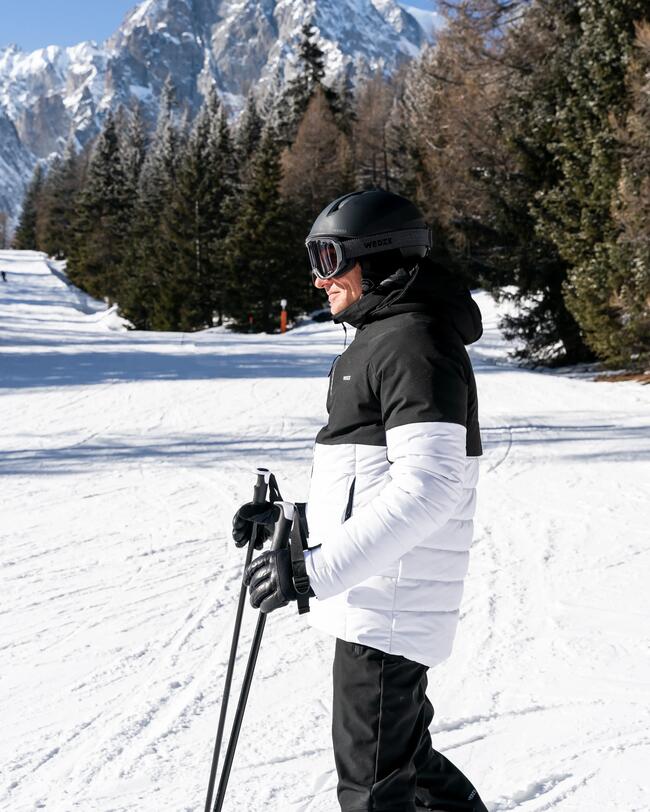 Ein Mann mit schwarz-weißer Skijacke, schwarzen Handschuhen, Skihelm und Stöcken steht auf einer Skipiste.