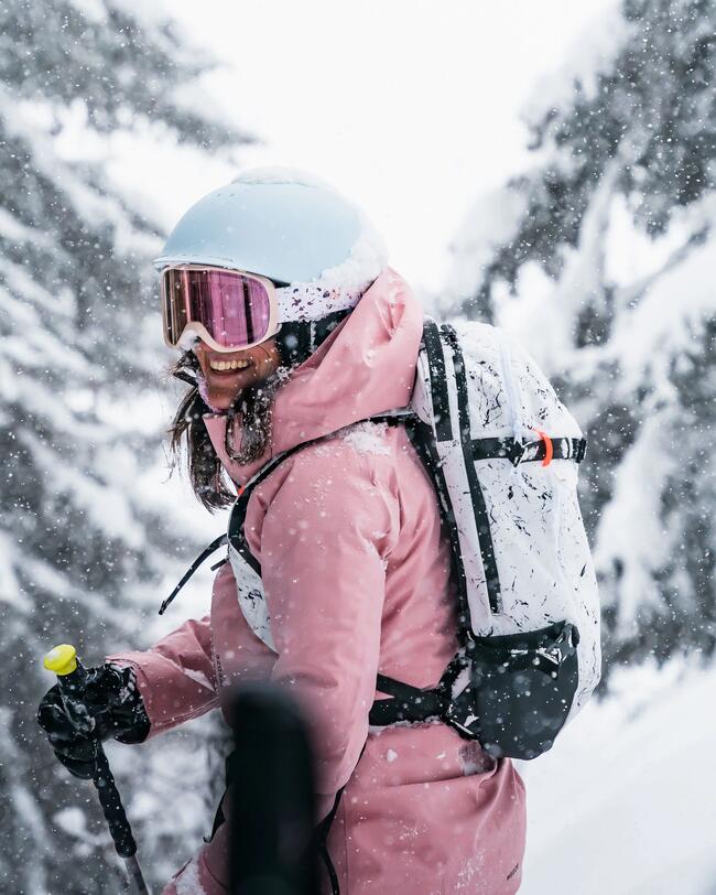 Eine Frau mit pinker Skijacke , weißem Skihelm und weißem Rucksack steht in einer verschneiten Winterlandschaft.