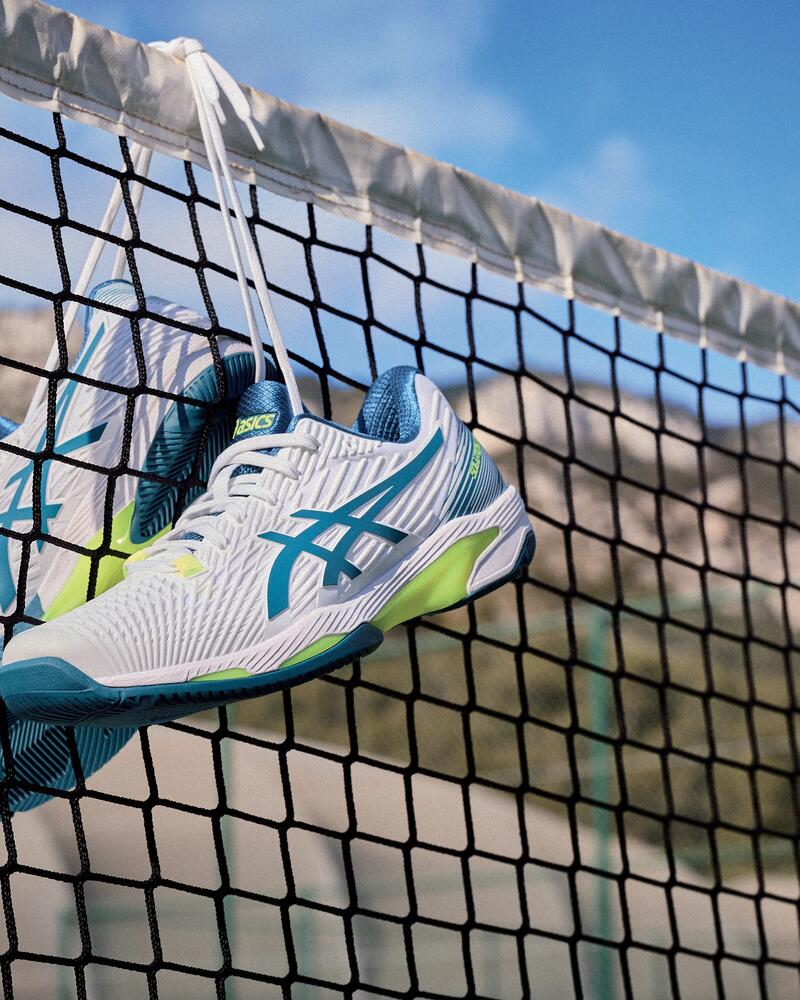 Ein Paar weiß-türkisfarbene „Asics“-Tennisschuhe mit neongelben Akzenten hängt an den Schnürsenkeln an einem Tennisnetz vor einem sonnigen Outdoor-Hintergrund.