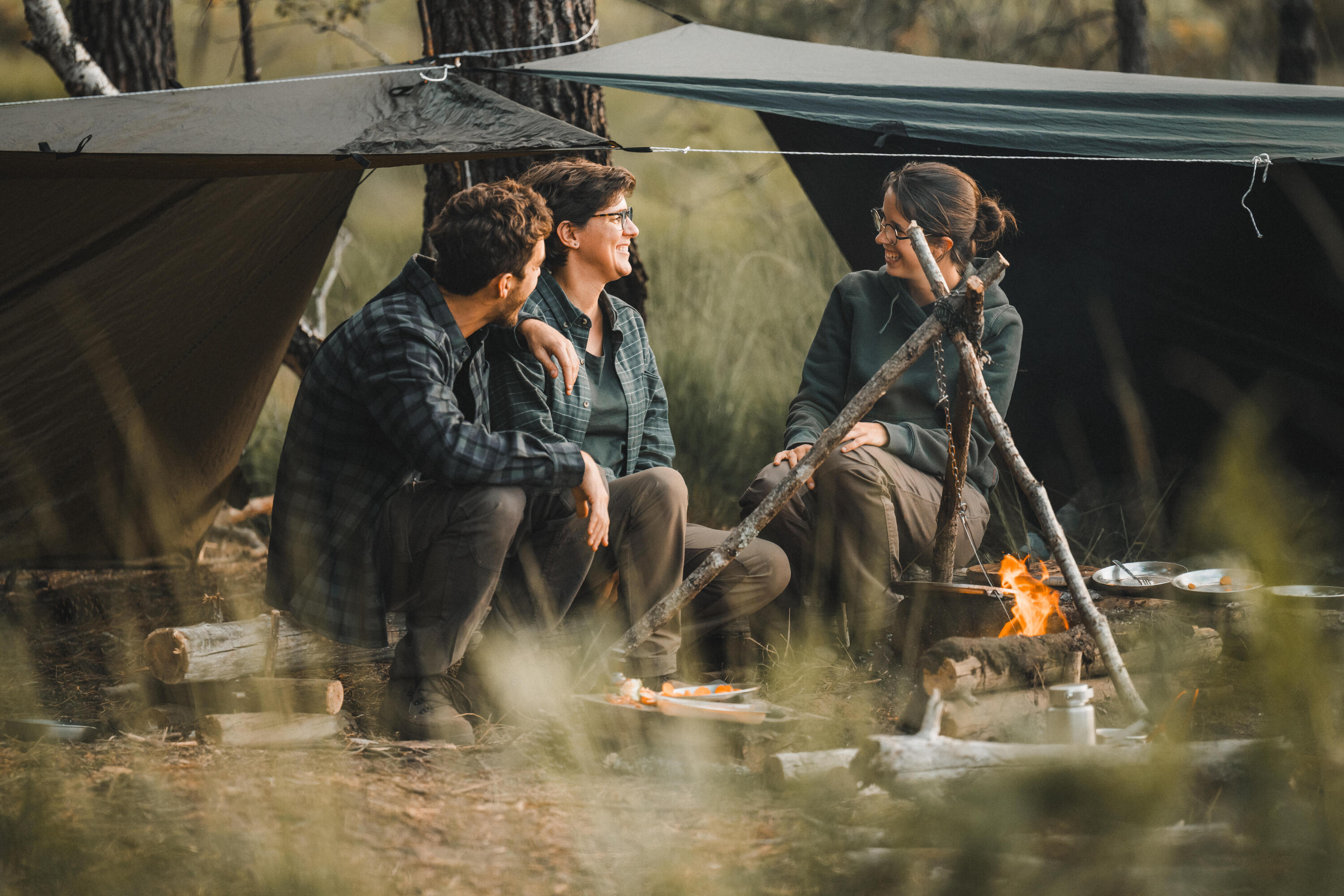 3 Personen sitzen am Lagerfeuer vor ihren Tarps und tragen Bushcraft Bekleidung