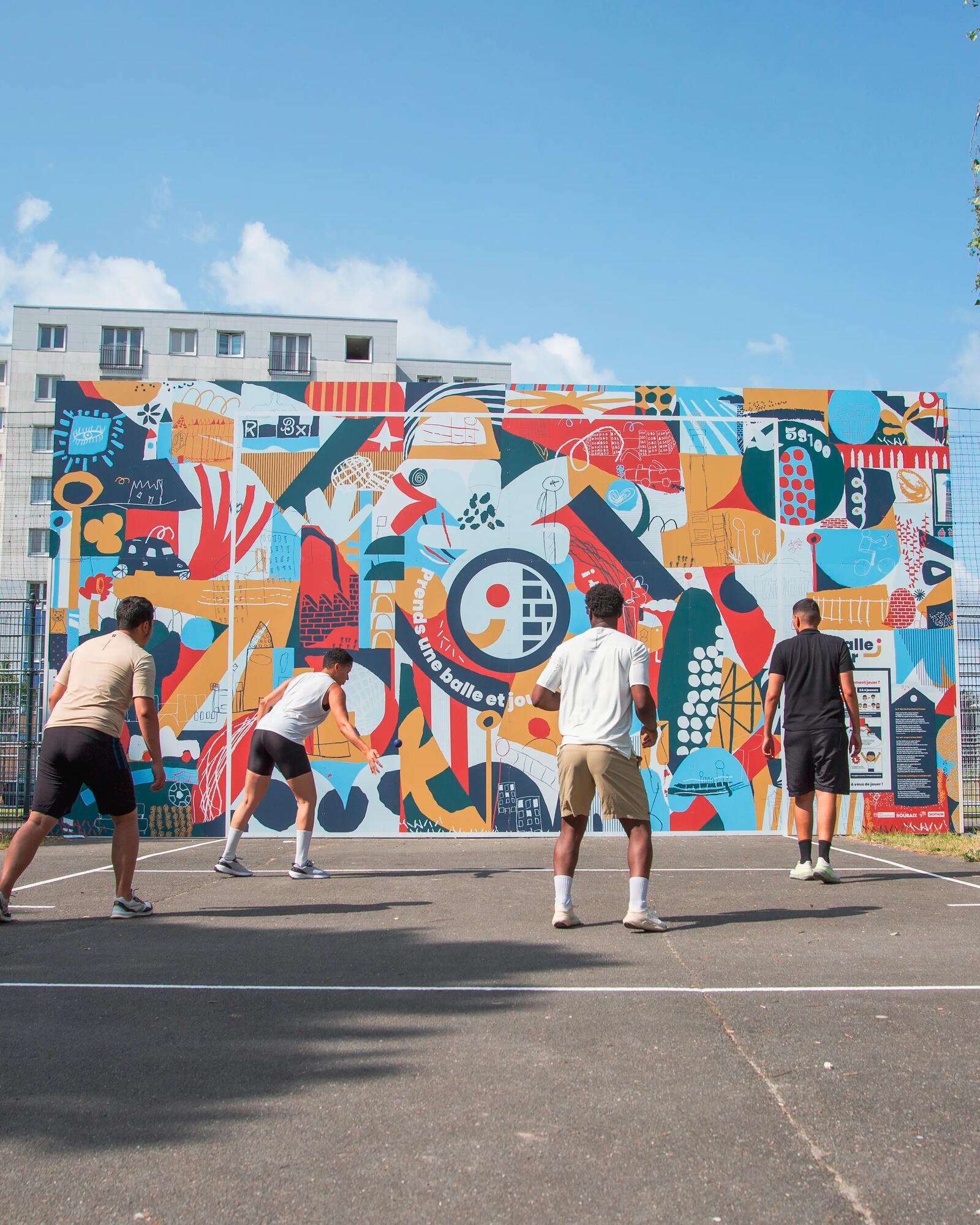Vier Sportler spielen auf einem urbanen Outdoor-Court Pelota gegen eine großflächige, bunt bemalte Wand mit abstrakten Motiven.