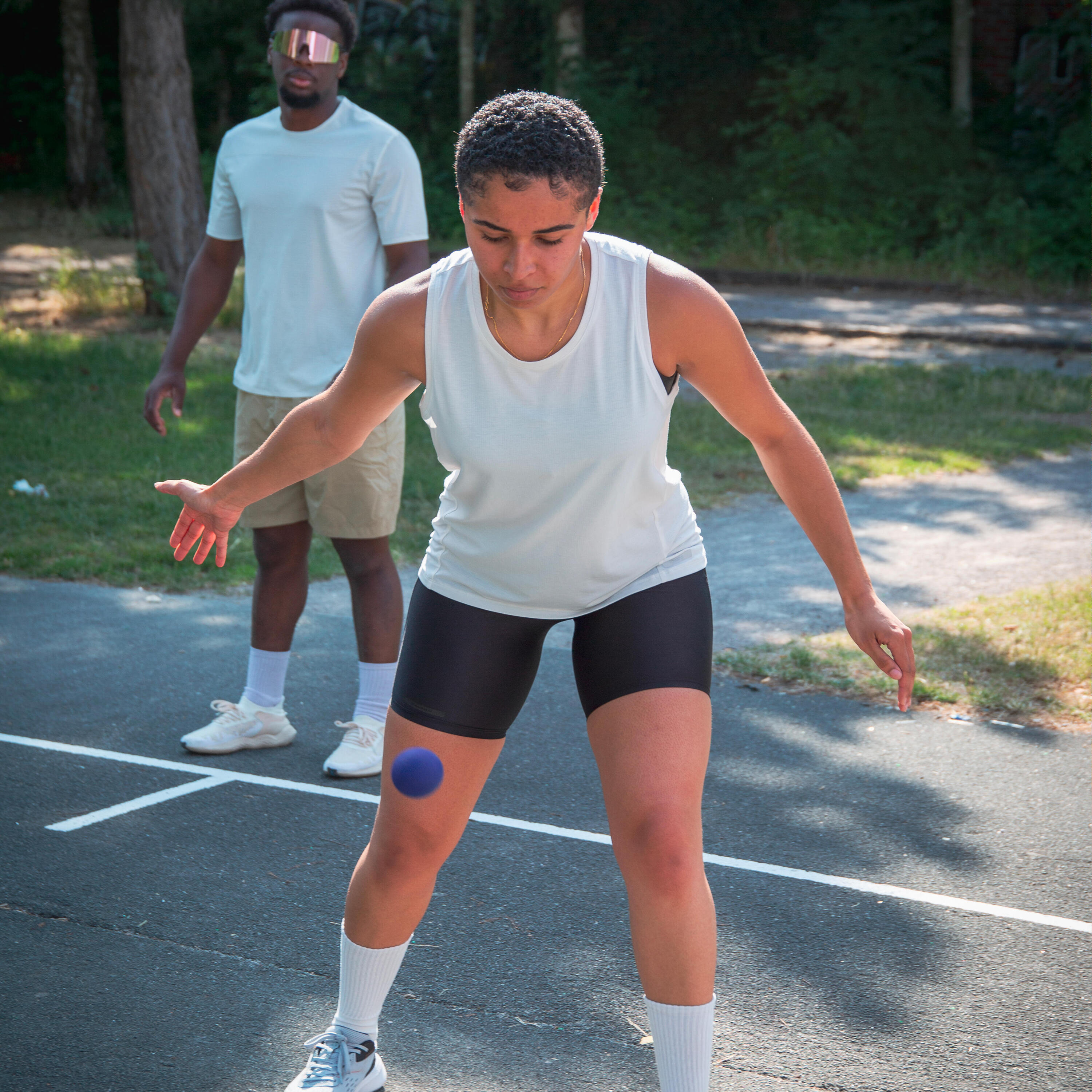 Junge Frau im weißen Sport-Top und schwarzer Radlerhose kickt einen kleinen blauen Ball auf einem Outdoor-Sportplatz; ein Mann im hellen Trainingsoutfit beobachtet sie im Hintergrund.