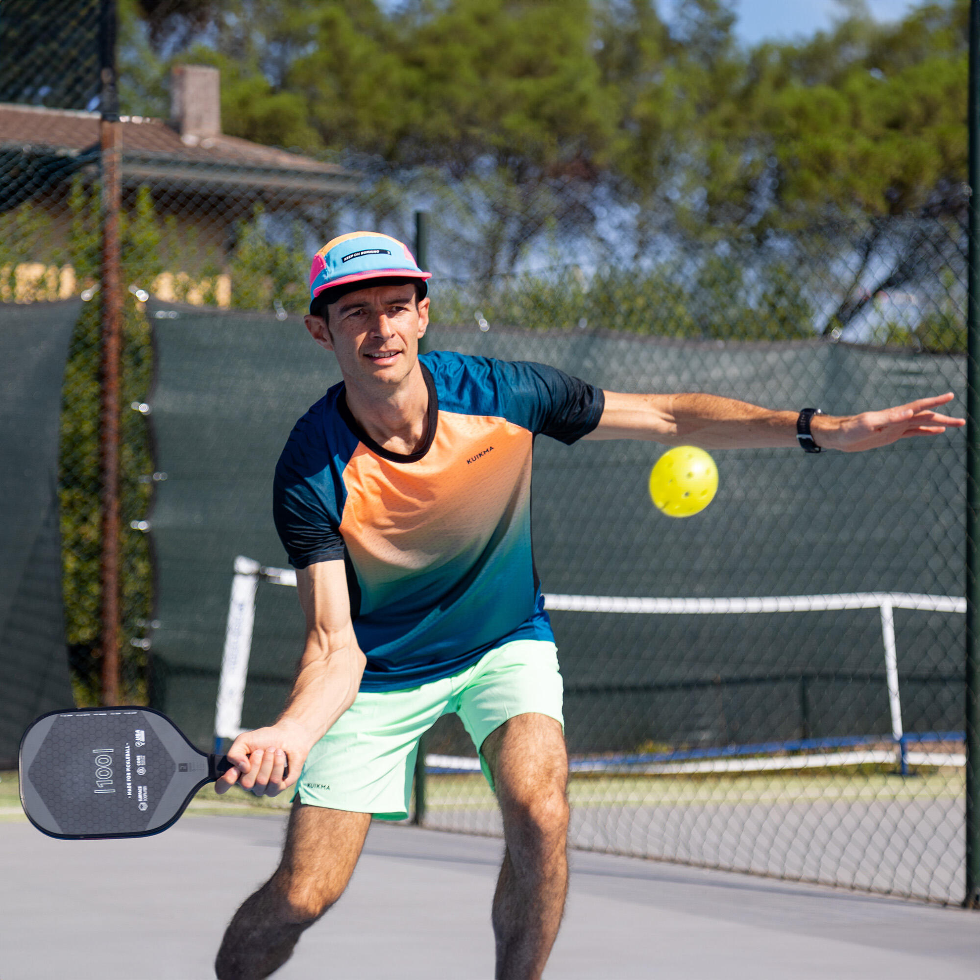 Pickleball-Spieler im blau-orangefarbenen Kuikma-Funktionsshirt, hellgrünen Shorts und bunter Cap holt mit einem schwarzen Kuikma-Pickleball-Schläger aus, um einen fliegenden gelben Ball zu schlagen.