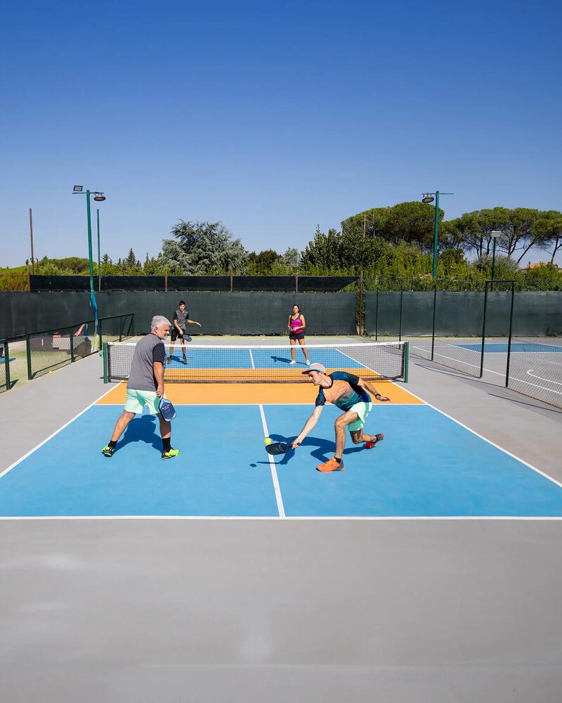 Vier Personen spielen Pickleball auf einem blau-orangenen Outdoor-Court, wobei ein Spieler im Vordergrund tief in die Knie geht, um den Ball mit seinem Paddel kurz vor dem Boden zu erreichen.