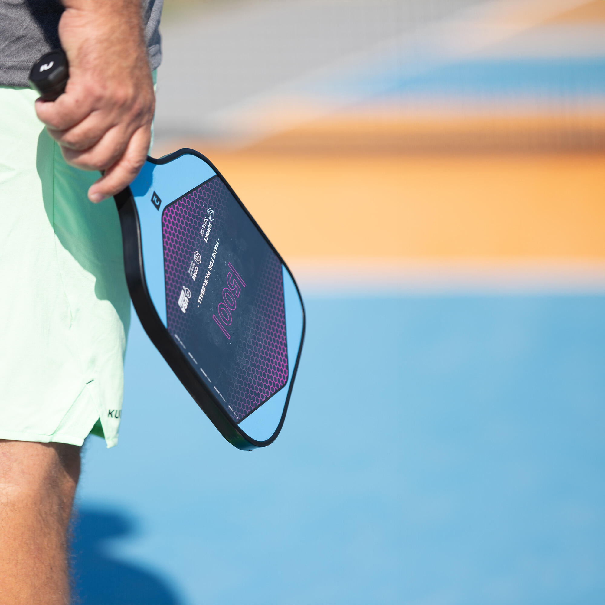 Nahaufnahme eines Pickleball-Spielers, der einen hellblauen Pickleball-Schläger mit schwarzem Rahmen und pink-violettem Waben-Design auf einem blauen Sportplatz hält.