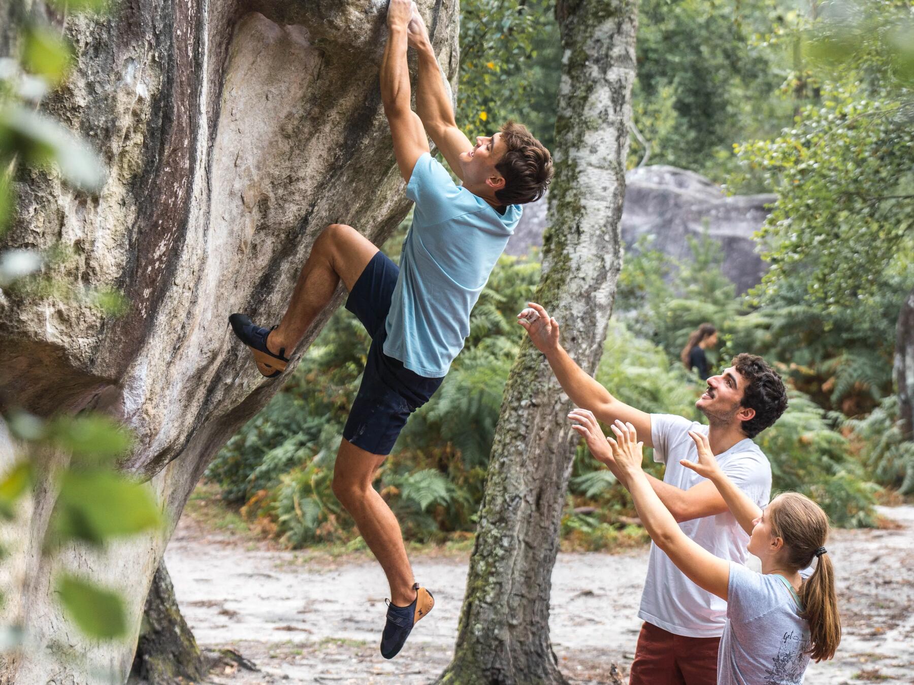 Mężczyzna w butach do wspinaczki uprawiający bouldering z asekuracją znajomych stojących pod skałą 