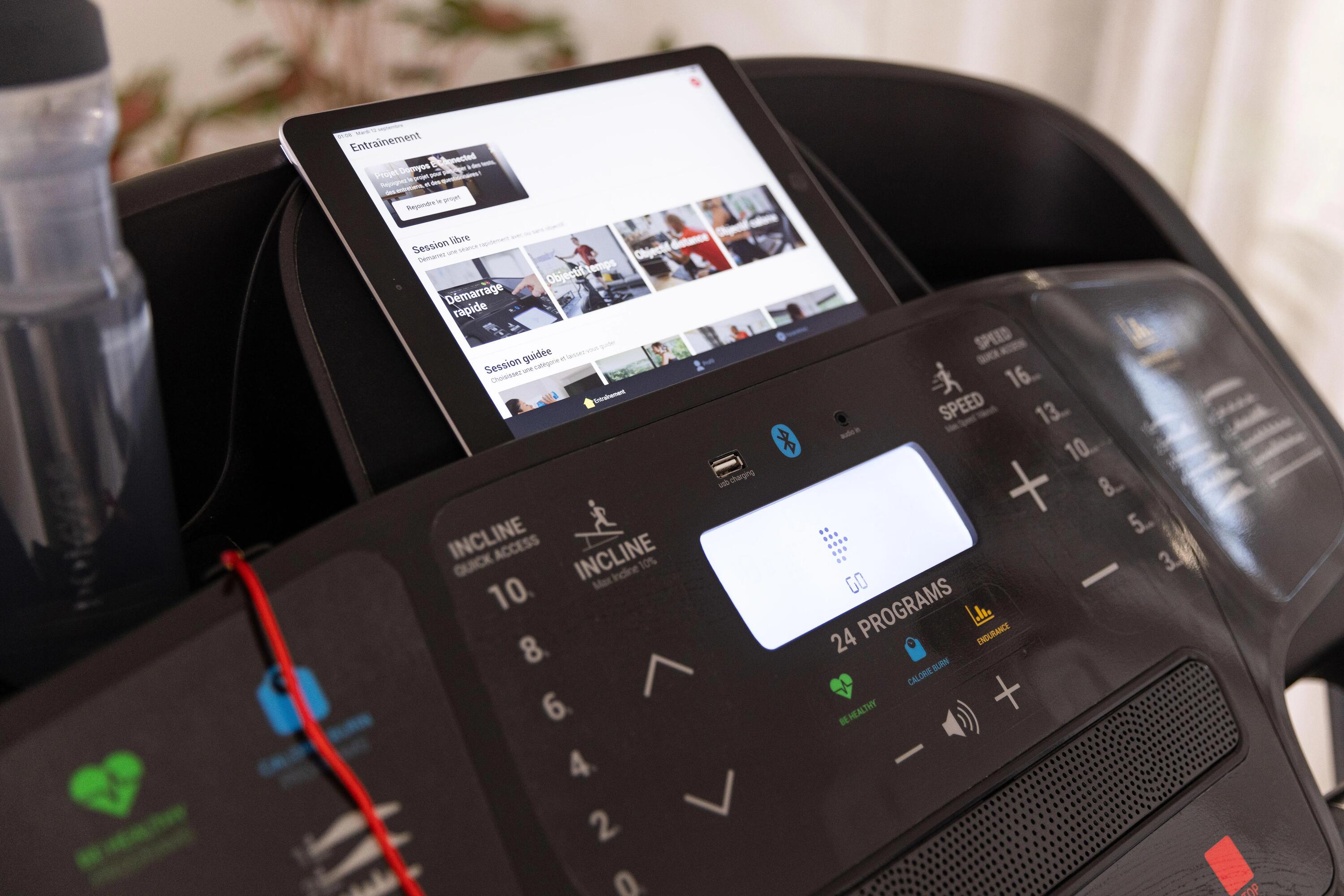 Bedienfeld eines Laufbands mit Knöpfen, kleinem Display und einem Tablet in der Halterung, das Trainingsprogramme zeigt.