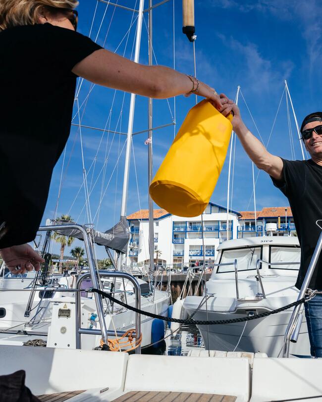Zwei Menschen auf einem Boot, übergeben einen gelben wasserdichten Sack, klarer Himmel im Hintergrund.