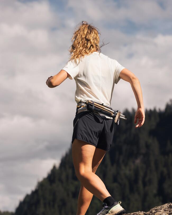 Person wandert auf felsigem Berg, trägt weißes T-Shirt und schwarze Shorts, bewaldete Hügel im Hintergrund, wolkiger Himmel.