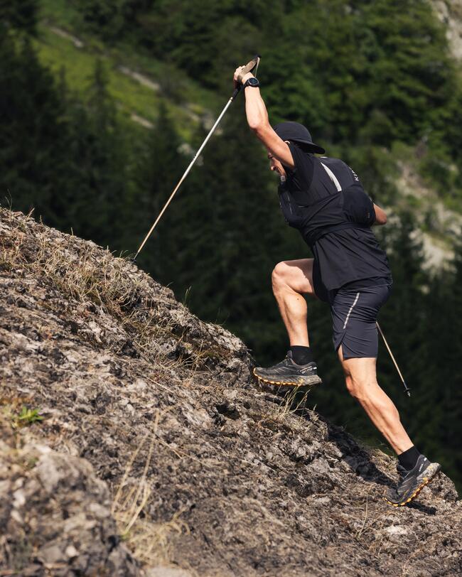 Person erklimmt felsigen Abhang mit Wanderstöcken, trägt schwarze Kleidung, Hintergrund aus grünem Wald und Felsen.