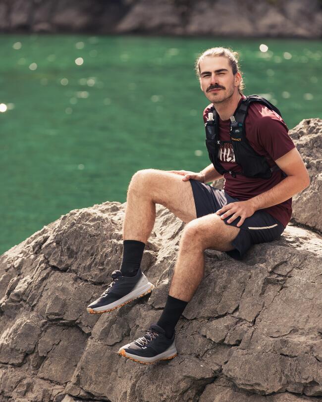 Mann mit dunklem Shirt und schwarzem Rucksack sitzt auf grauem Felsen vor grünem Wasser.