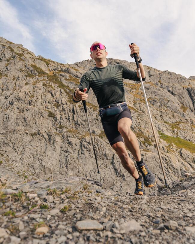 Mann mit Trekkingstöcken läuft über steinigen Pfad vor imposanten Felsen; trägt Sonnenbrille, dunkle Kleidung.