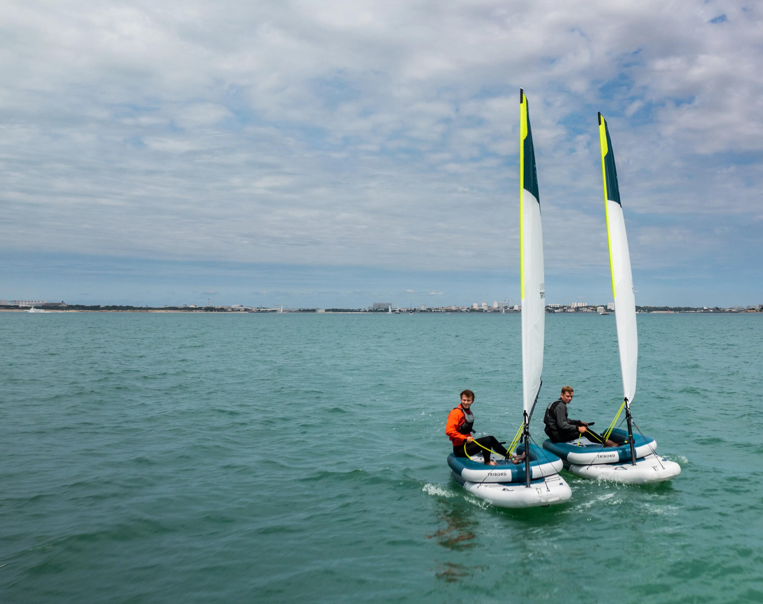 Eine weite Aufnahme zeigt zwei kleine Segelboote mit weißen und neongrünen Segeln, die mit jeweils einer Person besetzt auf ruhigem, türkisblauem Wasser unter leicht bewölktem Himmel segeln.