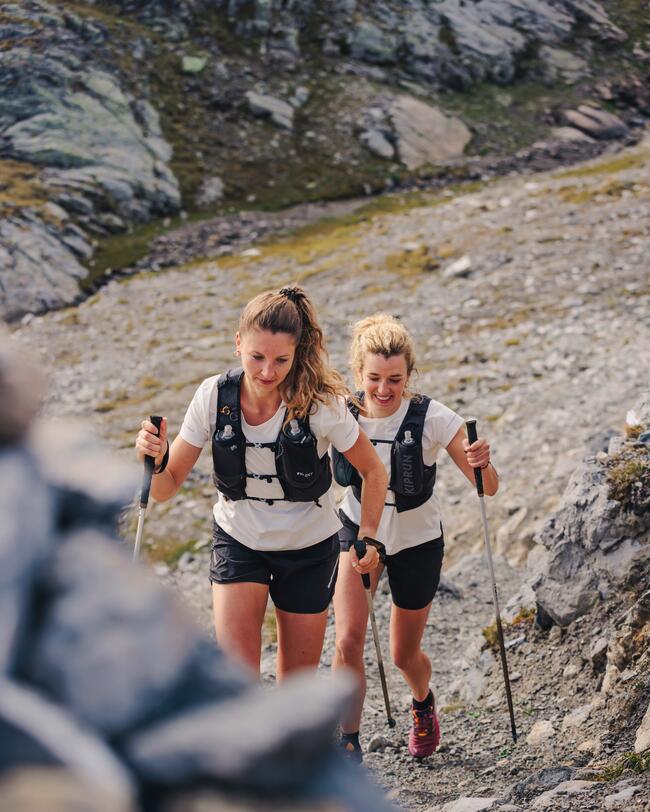 Zwei wandernde Frauen in heller Sportkleidung auf felsigem Bergpfad, umgeben von grauen Steinen und spärlicher Vegetation.