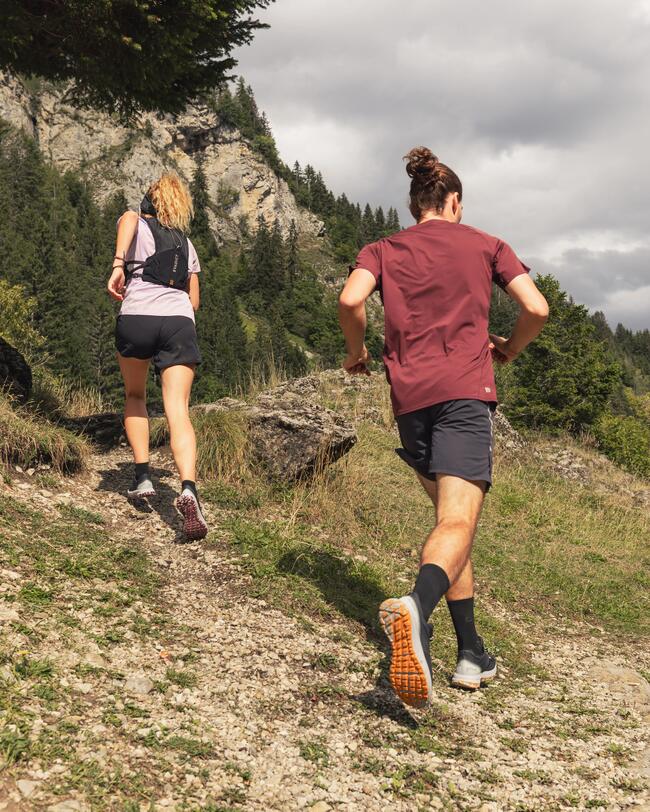 Zwei Personen joggen bergauf auf einem steinigen Pfad, umgeben von grünen Bäumen und grauen Felsen, unter bewölktem Himmel.