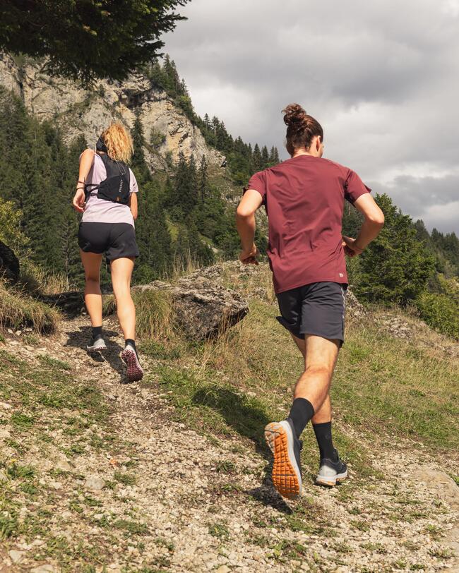 Zwei Menschen joggen einen steinigen Waldpfad hinauf, umgeben von grünen Bäumen, einem klaren Himmel und Felsformationen.