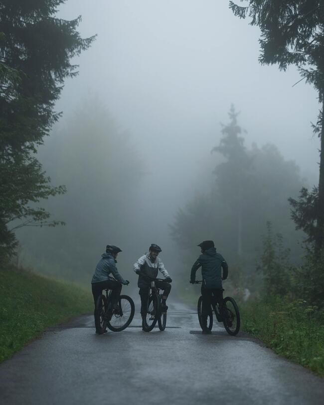Drei Radfahrer stehen mit ihren Mountainbikes auf einer asphaltierten Straße in einem dichten, nebligen Wald. Die Szene ist in gedämpften Grau- und Grüntönen gehalten, wobei die Silhouetten der Bäume im Hintergrund im Nebel verschwimmen.