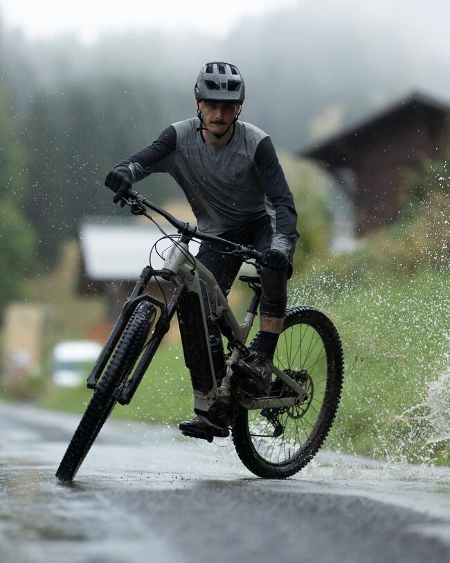 Eine Fotografie zeigt zwei Mountainbiker mit Helmen auf einer nassen Asphaltstraße vor einer nebligen Berg- und Kulissenlandschaft. Der vordere Radfahrer fährt dynamisch durch eine Pfütze, wodurch Wasser seitlich hochspritzt, während die zweite Person mit etwas Abstand dahinter fährt.