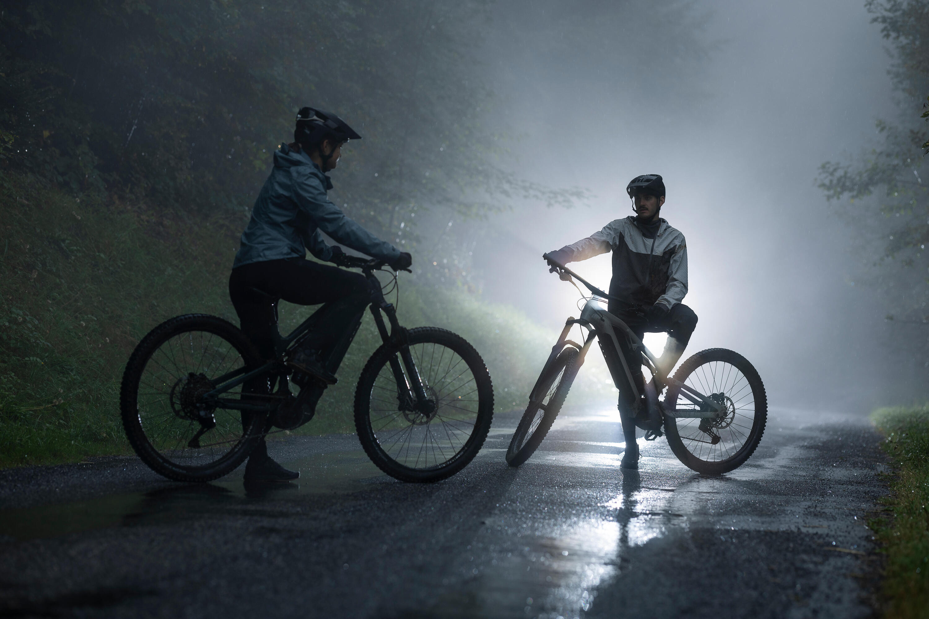 Zwei Mountainbiker mit Rockrider Feel Helmen und Regenbekleidung stehen bei Nebel und nasser Fahrbahn auf einer Bergstraße.
