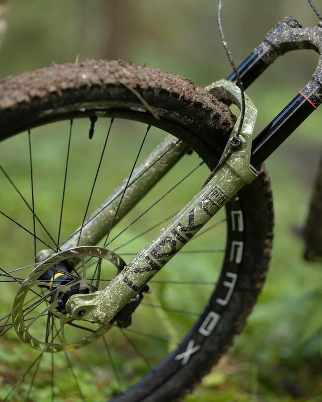 Grünes, schlammverschmiertes Mountainbike-Vorderrad mit grobem Reifenprofil und silberner Bremsscheibe im grünen Wald.