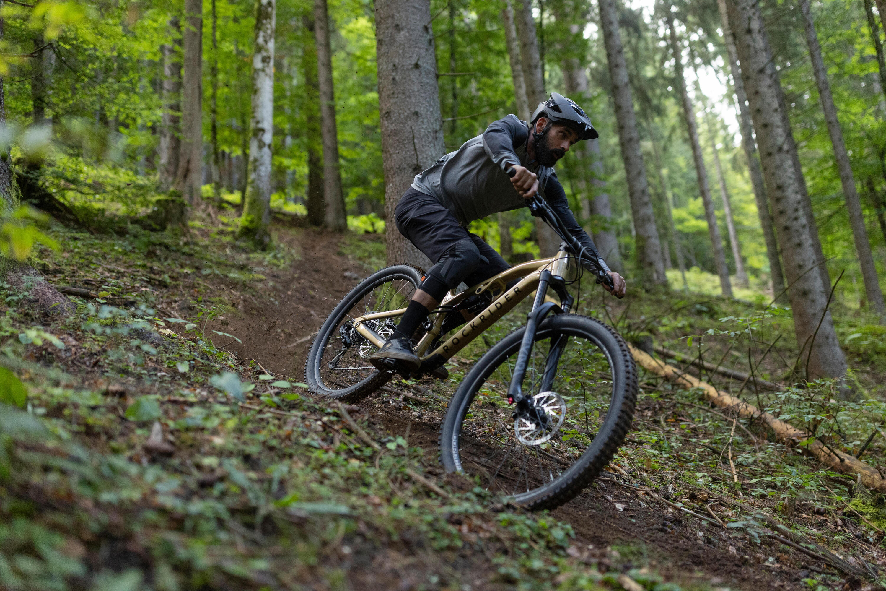 Mountainbiker fährt eine steile Kurve auf einem Wald-Trail mit dem goldenen Rockrider FEEL 700 LT Mountainbike Fully.
