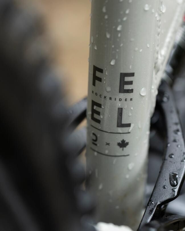 Eine Nahaufnahme des hellgrauen, mit Wassertropfen bedeckten Rahmens eines Mountainbikes, auf dem vertikal der schwarze Schriftzug „FEEL“ sowie „ROCKRIDER“ und ein Ahornblatt-Symbol zu sehen sind. Der Hintergrund und die umliegenden Fahrradteile sind unscharf abgebildet.