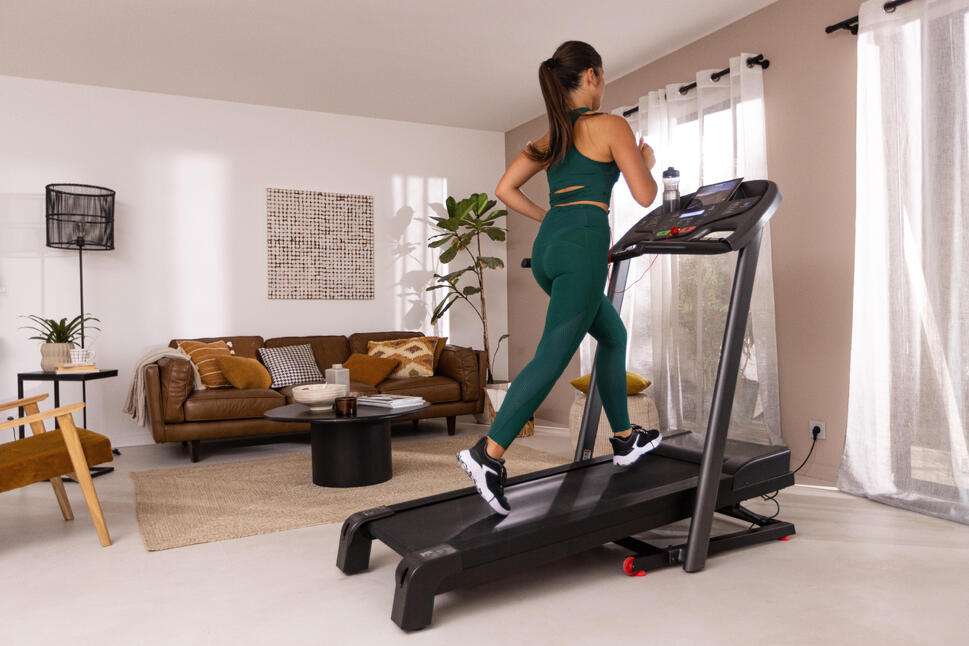 Ženska v temno zelenem športnem kompletu teče na črni tekalni stezi Domyos T520B v dnevni sobi.