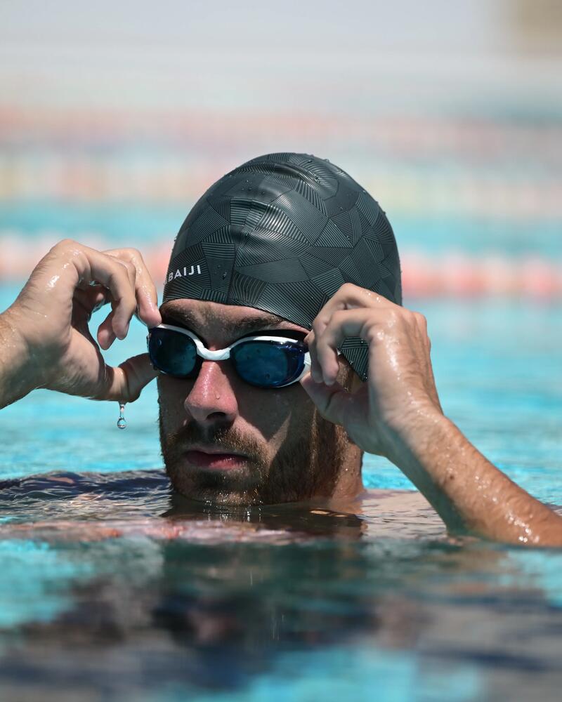 Mann mit schwarzer Badekappe im blauen Wasser, setzt schwarze Schwimmbrille auf, Lichtreflexionen auf der Wasseroberfläche.