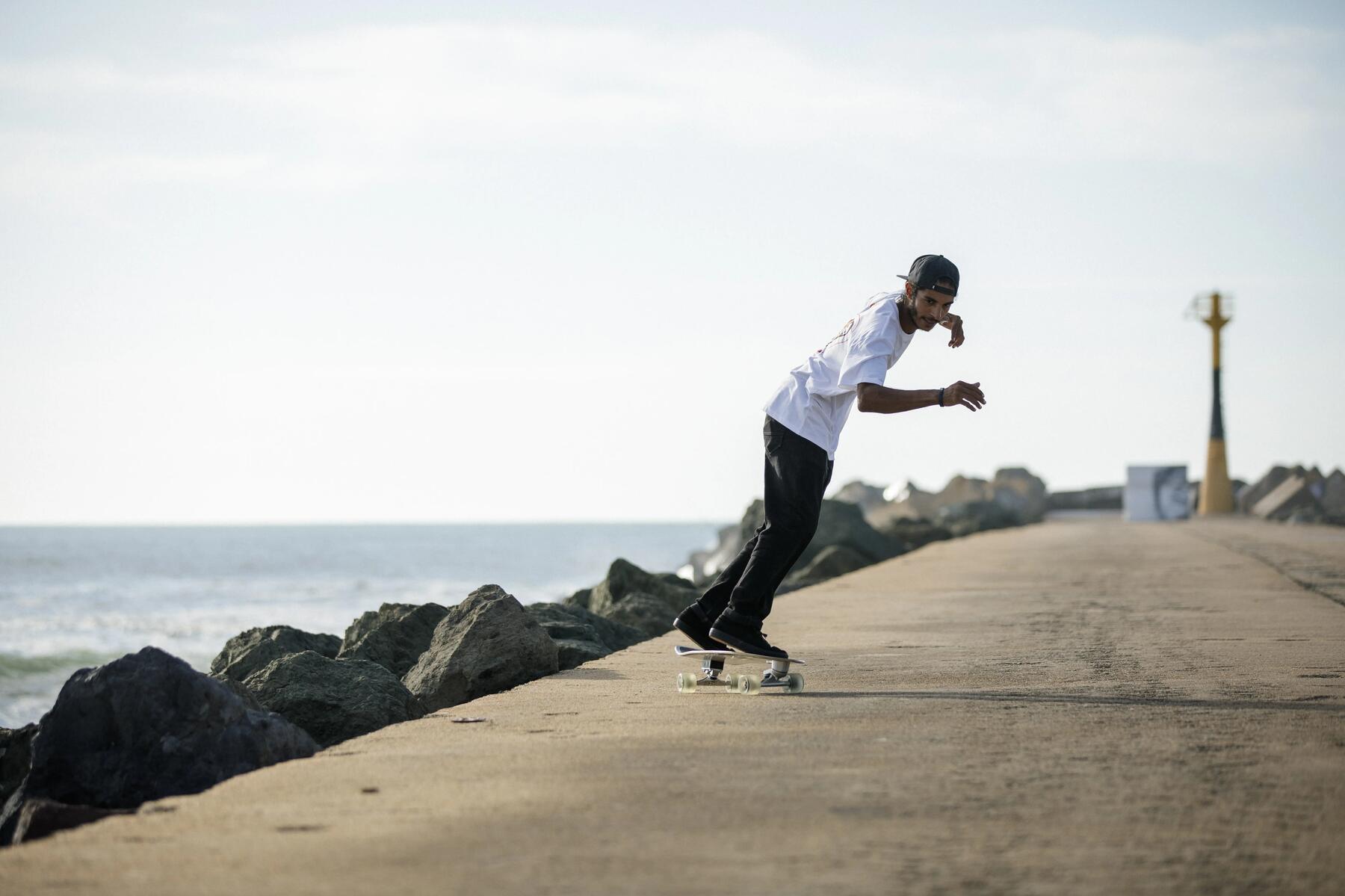 Deskorolkowiec w czapce z daszkiem jadący na desce typu longboard po utwardzonej nawierzchni w pobliżu wody
