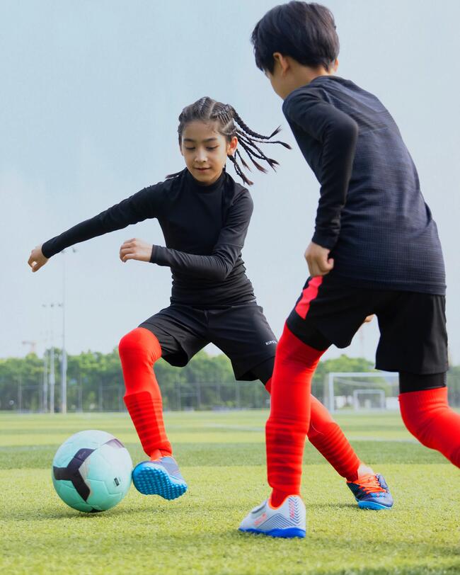 Zwei Jungs im schwarzen Keepdry Shirt und roten Stutzen spielen Fußball. Über ihnen blauer Himmel und Bäume verschwommen im Hintergrund.