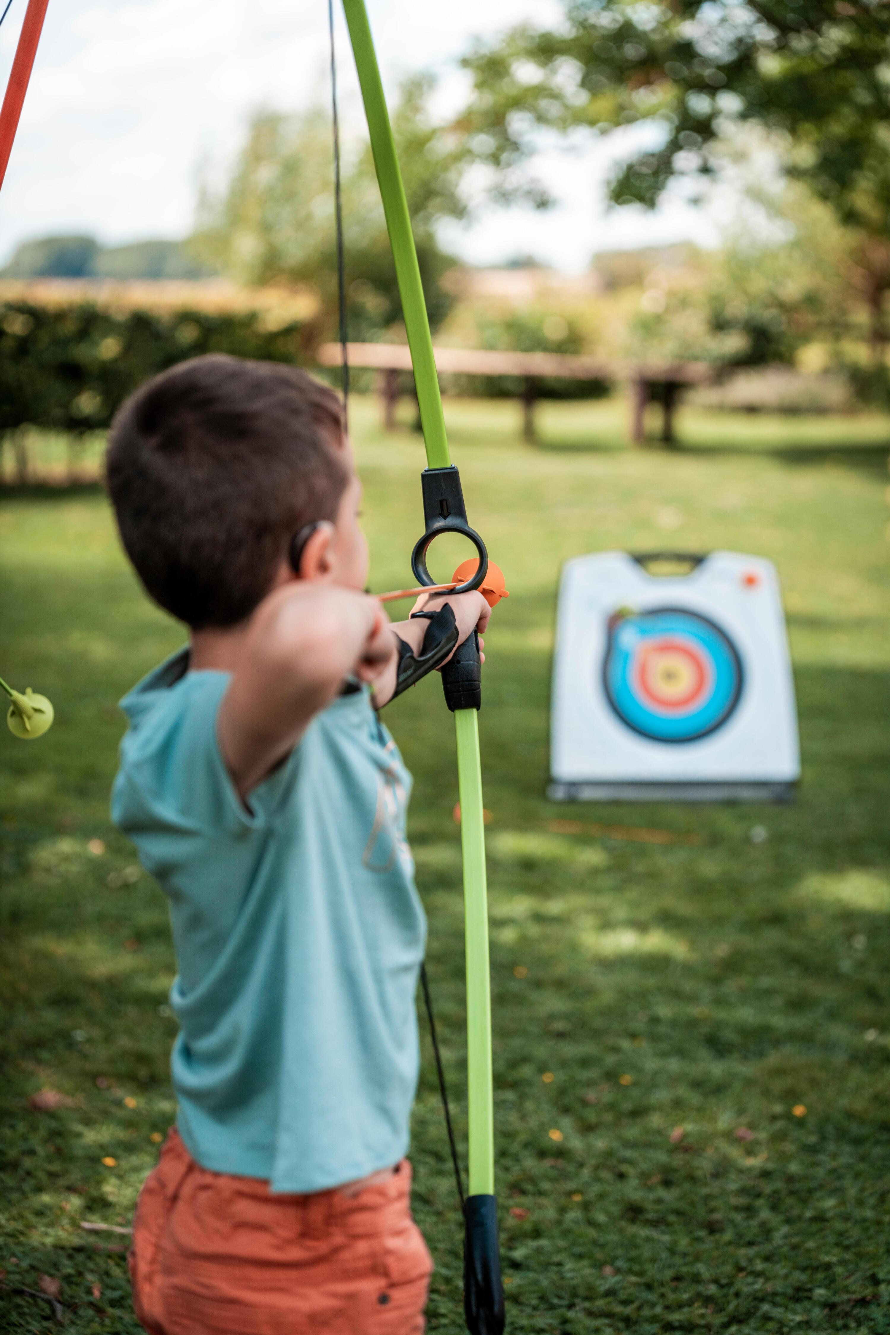 Nahaufnahme eines Jungen mit Hörgerät von hinten, der mit einem grünen Spielzeug-Bogen auf eine farbige Zielscheibe im Garten zielt.