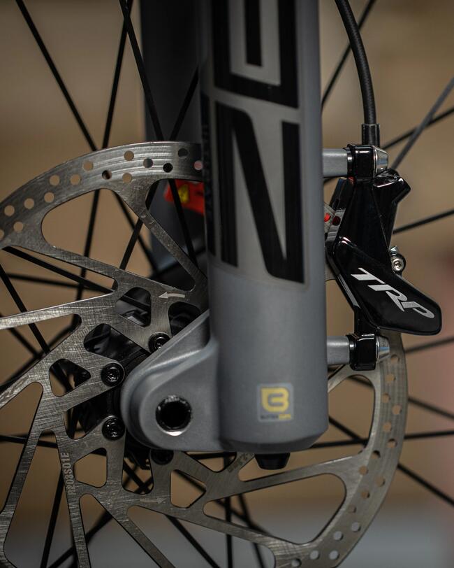 Graue Fahrradgabel mit schwarzem und silbernem Scheibenbremsen-Mechanismus, scharfe Linien und metallische Struktur.