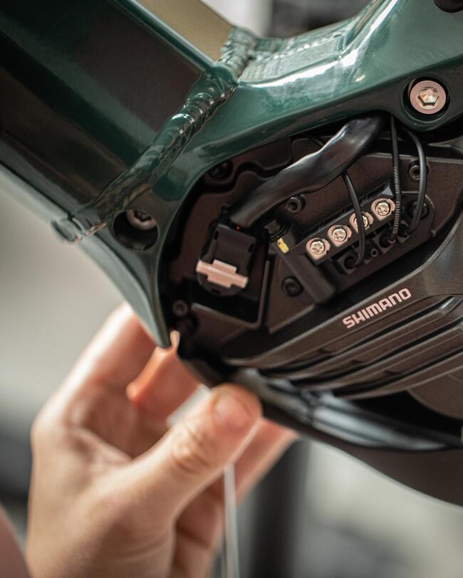 Schwarzer Shimano-E-Bike-Motor mit sichtbaren Kabeln an grünem Rahmen, glänzende und matte Oberflächen, Hand im Vordergrund.
