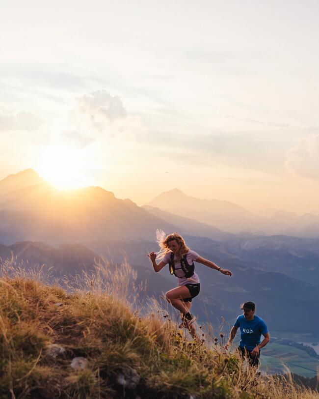 Zwei wandernde Personen bei Sonnenuntergang auf einem grasbewachsenen Berghang, leuchtender Himmel mit orange und rosa Tönen.