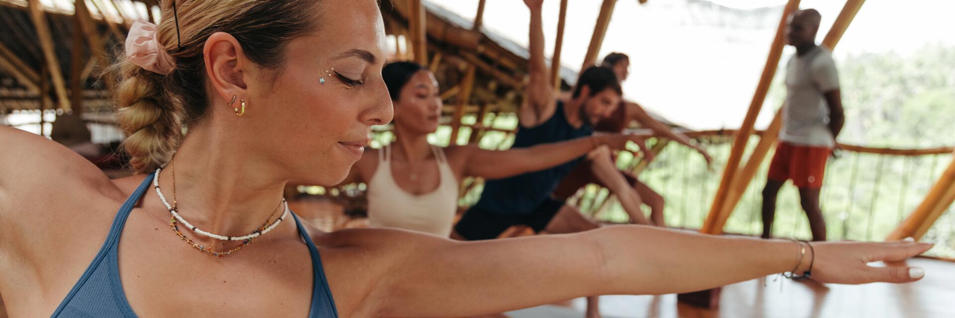 Personen bei Yoga Asanas. Frau im Vordergrund mit blauem Sport-BH, Personen im Hintergrund auf Yogamatten.