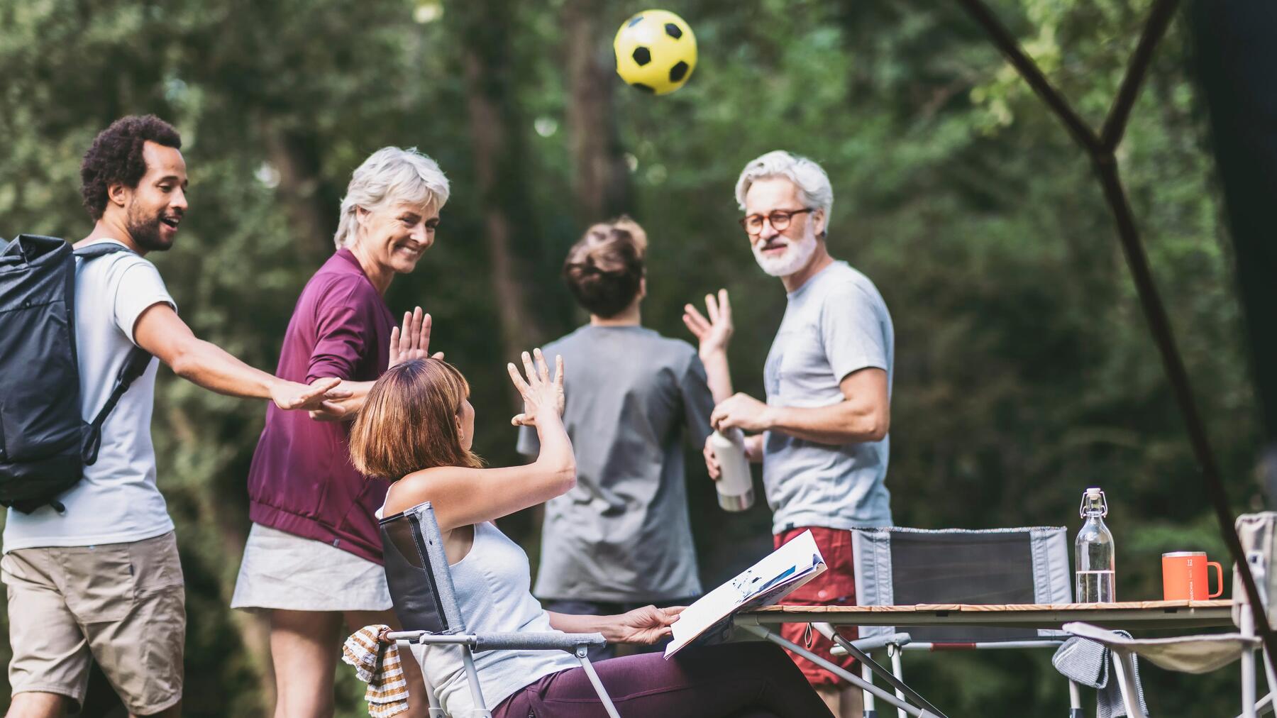 Familie spielt mit Ball, Frau sitzt am Campingtisch neben den Campingzelten.