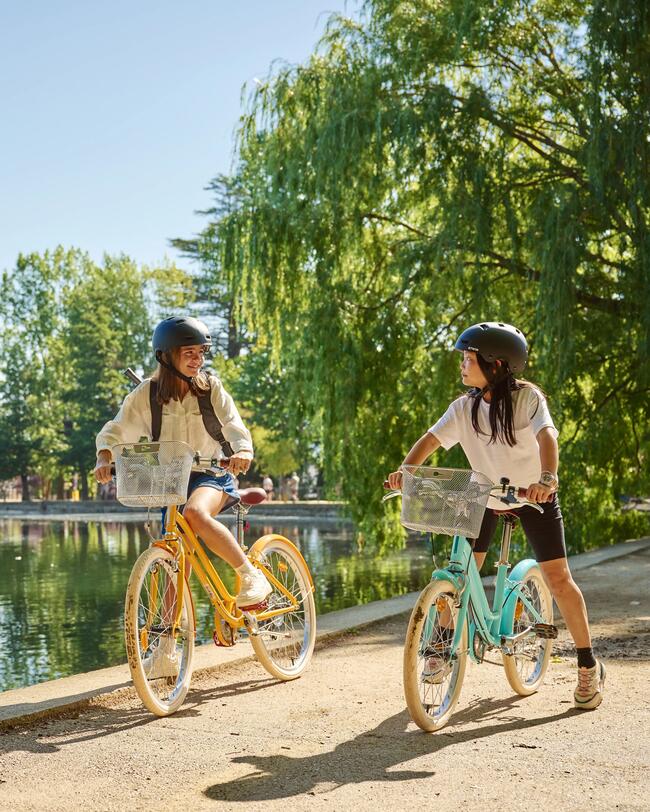 Zwei Kinder mit schwarzen Helmen stehen mit gelbem und türkisem Fahrrad im Park; im Hintergrund Teich und Bäume