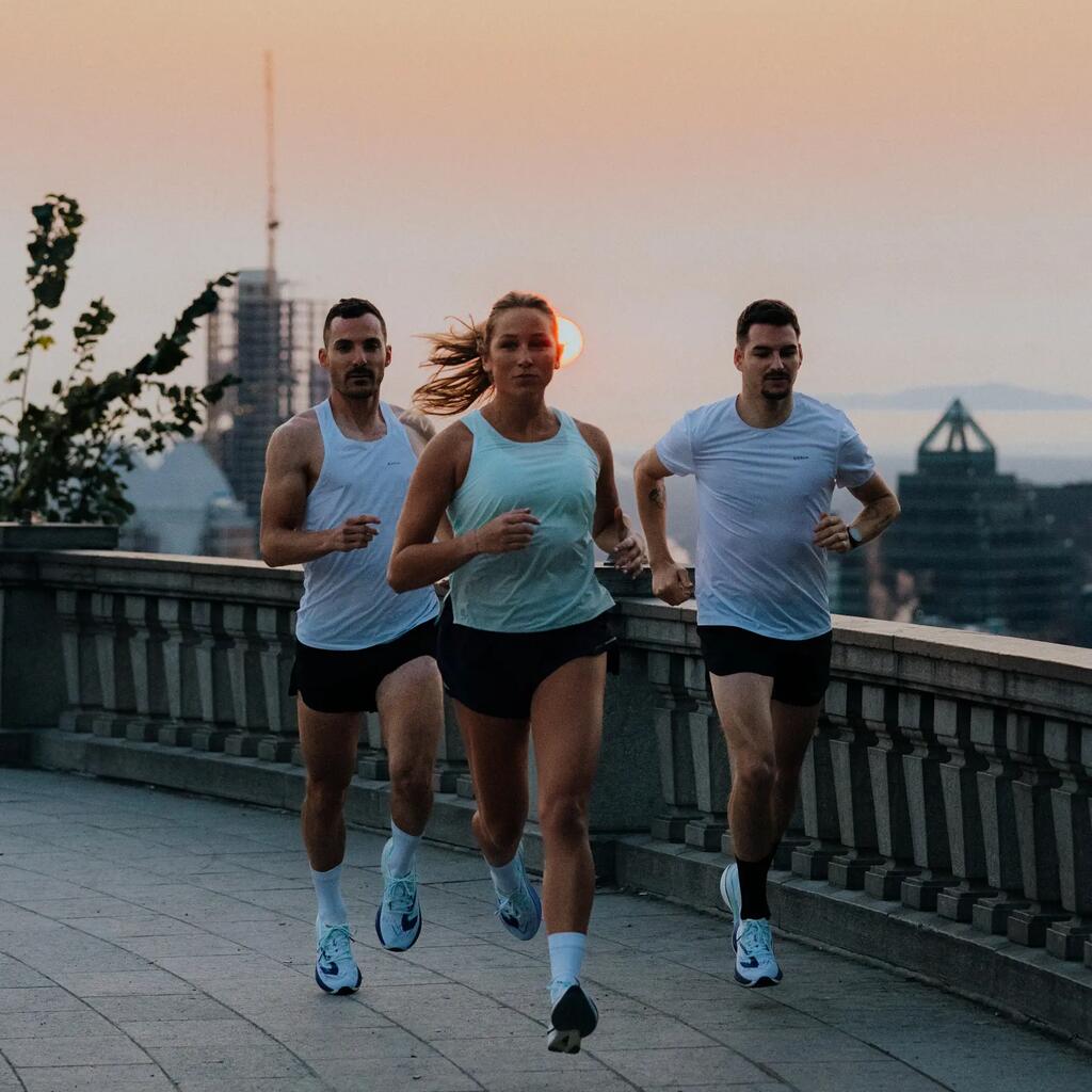 Trije tekači v športnih oblačilih Kiprun tečejo po tlakovani poti ob ograji razgledne točke.