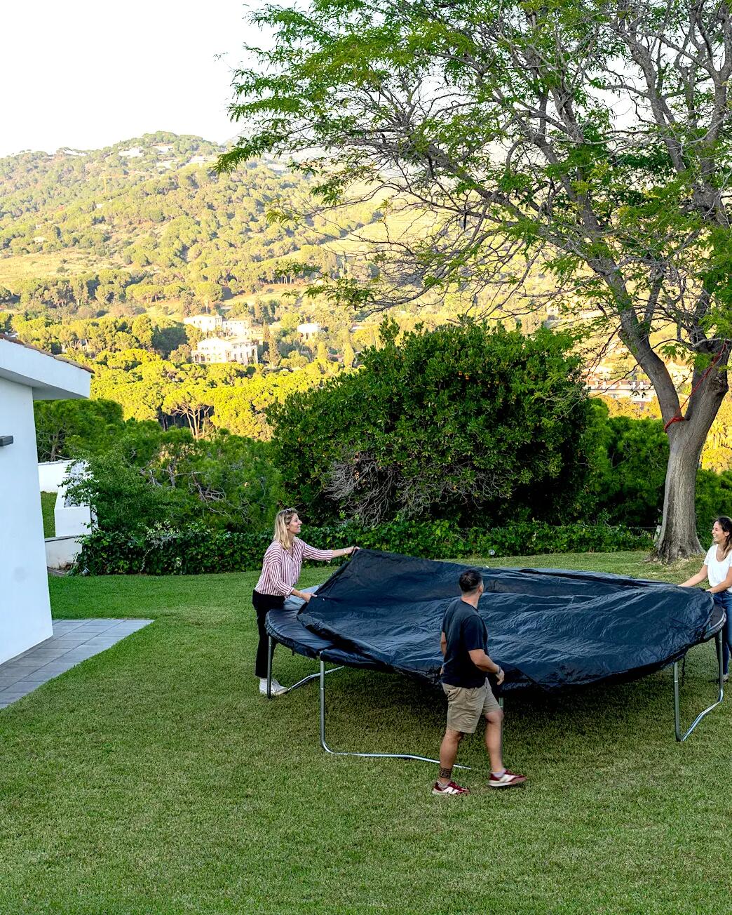 Drei Personen ziehen eine schwarze Schutzabdeckung über ein Trampolin auf einer Rasenfläche vor hügeliger Landschaft.