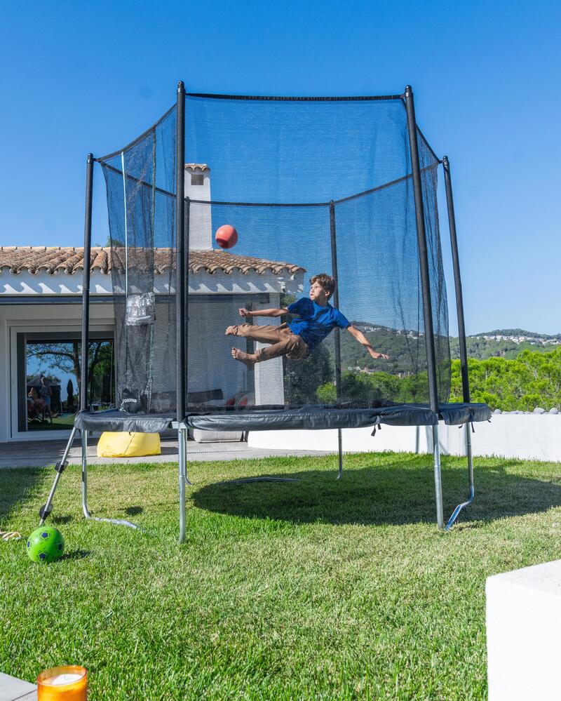 Ein Junge macht einen Seitfallzieher in einem Trampolin mit Fangnetz im sonnigen Garten vor einem weißen Haus.