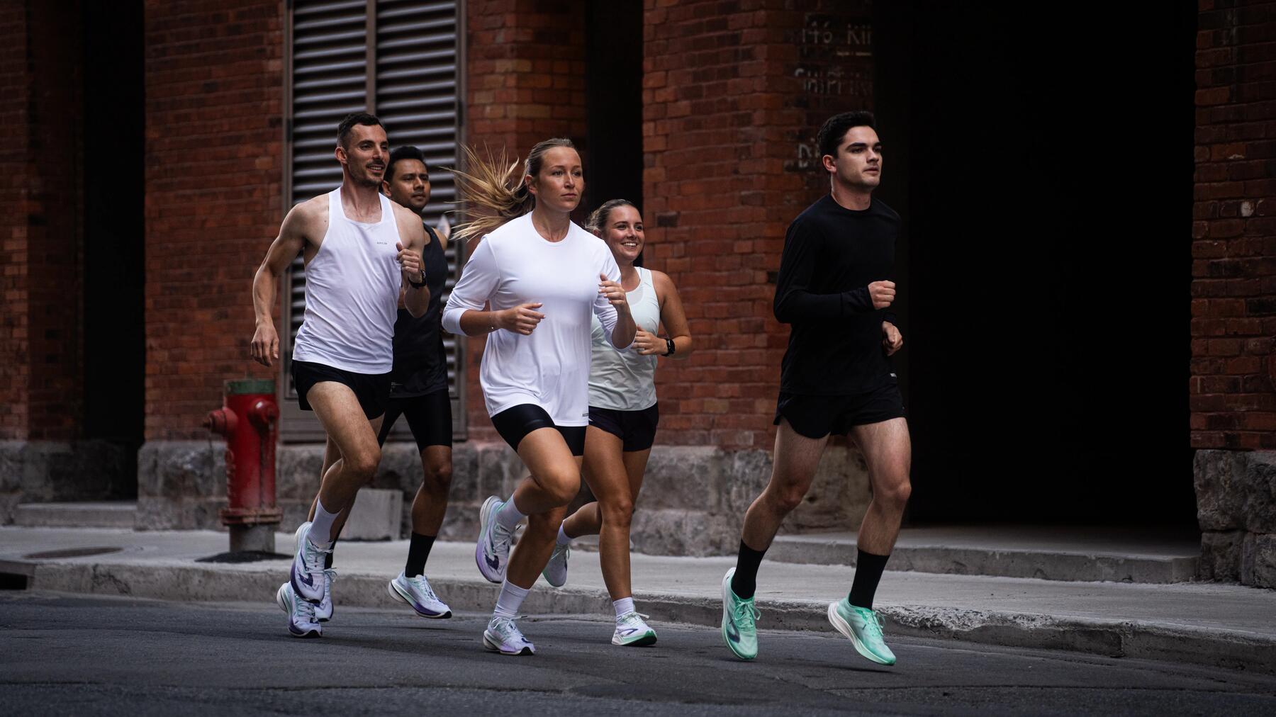Fünf sportliche Menschen joggen auf der Straße vor roten Backsteinwänden, tragen schwarze und weiße Sportkleidung.
