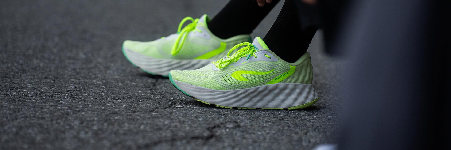 Neongelbe Laufschuhe mit weißen, dick profilierten Sohlen auf grauem Asphalt, daneben schwarze Hose und Socken.