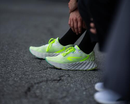 Neongelbe Laufschuhe mit weißen, dick profilierten Sohlen auf grauem Asphalt, daneben schwarze Hose und Socken.