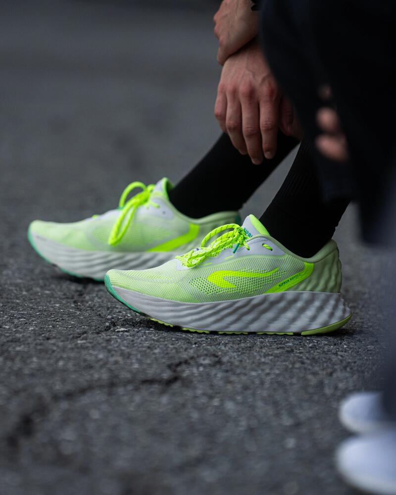 Neongelbe Laufschuhe mit weißen, dick profilierten Sohlen auf grauem Asphalt, daneben schwarze Hose und Socken.