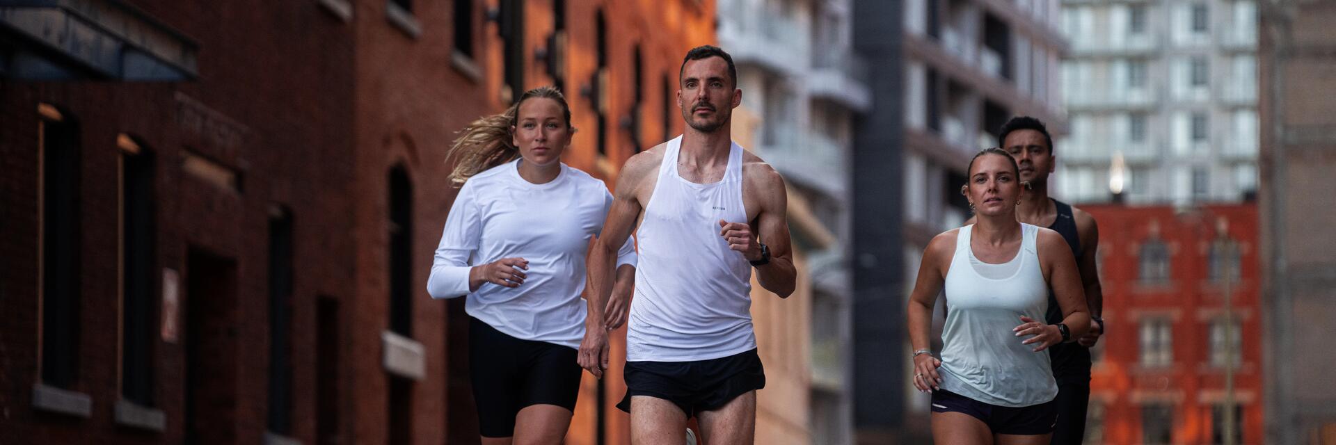 Vier Personen joggen in sportlicher Kleidung durch eine Stadt zwischen roten Ziegelhäusern und modernen Hochhäusern bei Tageslicht.