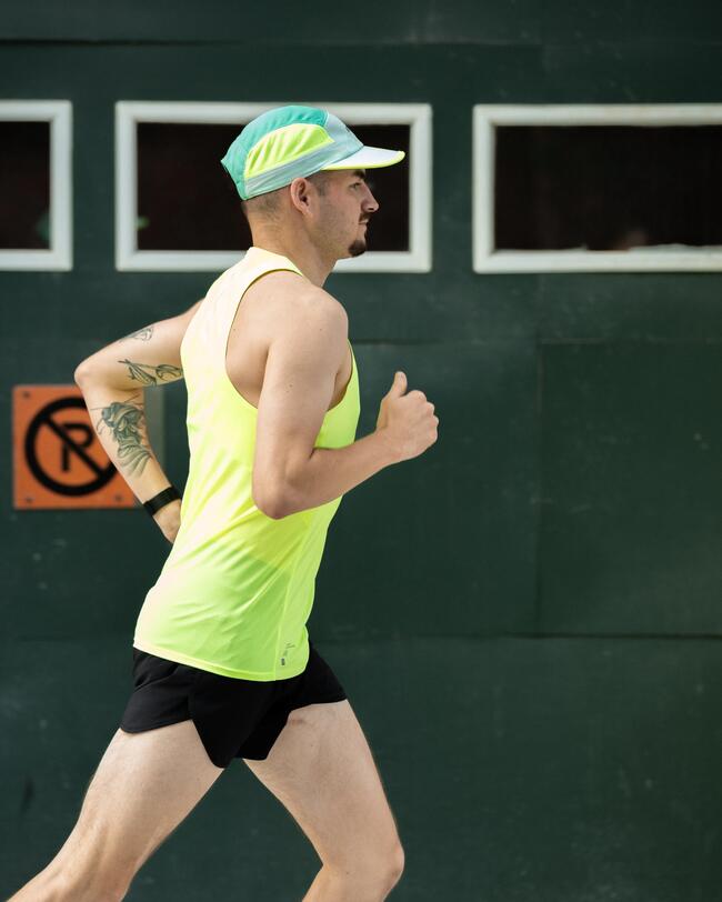 Mann in neonfarbenem Tanktop und Shorts beim Laufen vor grüner Wand mit gelbem Hut und Tätowierung.