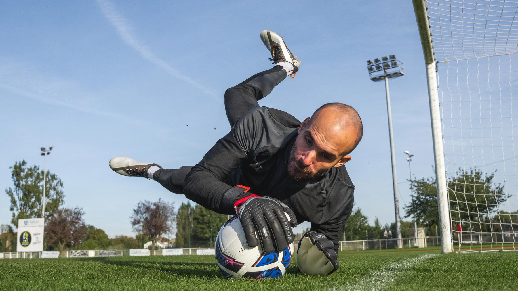 Ein glatzköpfiger Torwart in schwarzer Kleidung hechtet spektakulär durch die Luft und begräbt den Ball am Boden unter seinen Händen.