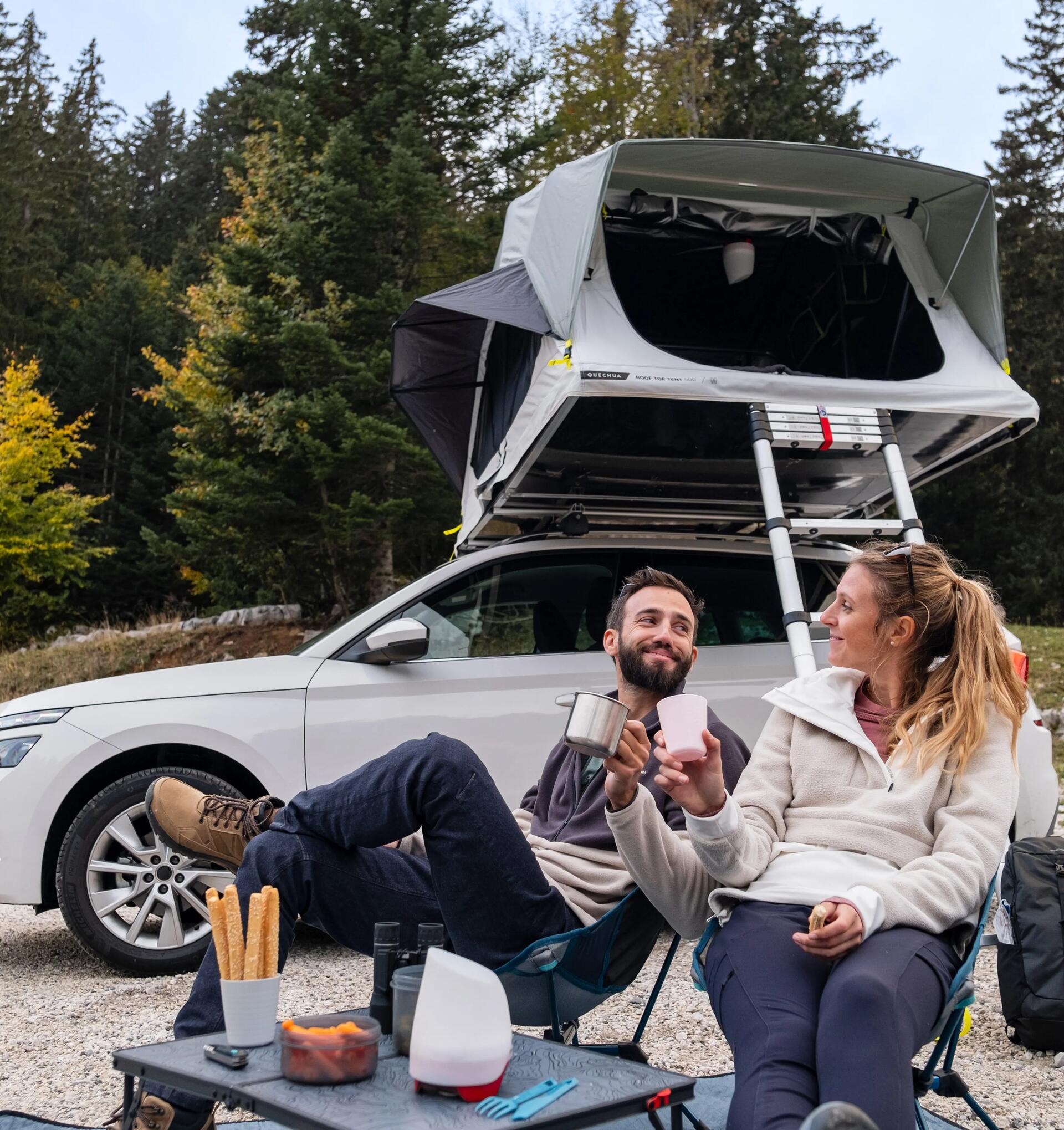 a couple sat outside in front of their car with a rooftop tent  set up on top