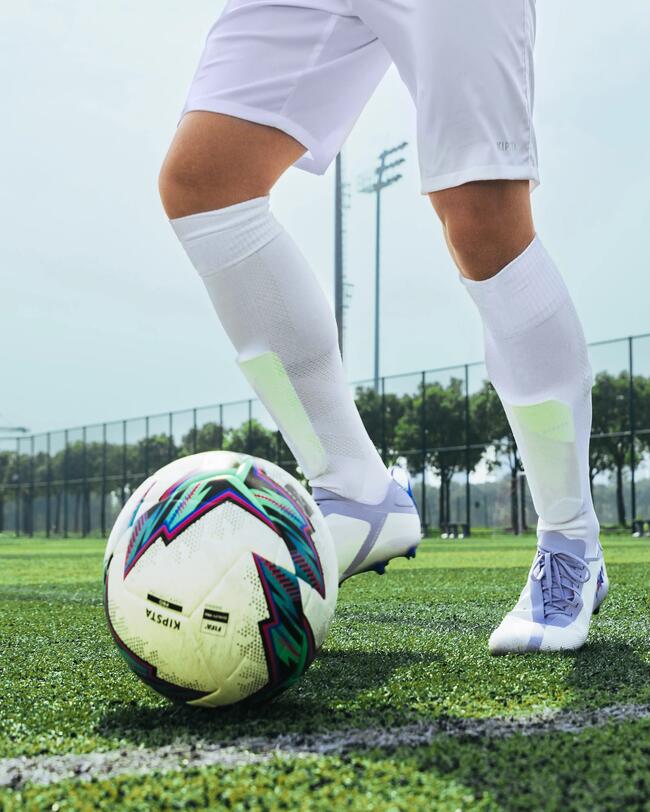 Person in weißem Fußball-Outfit nimmt Ball an. Bäume im Hintergrund, klarer Himmel.