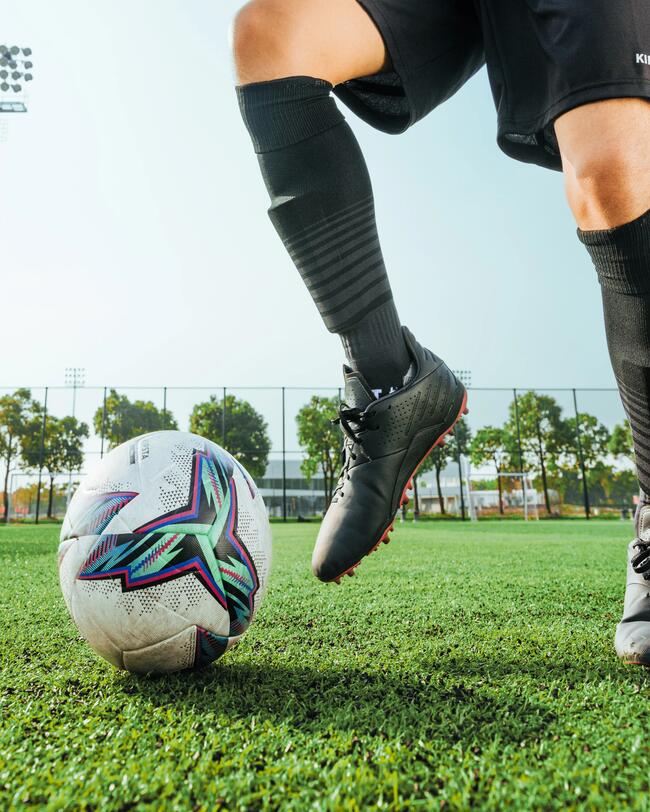 Person mit schwarzen Stutzen, Shorts und Schuhen dribbelt leicht verdreckten Ball. Bäume im Hintergrund, klarer Himmel.